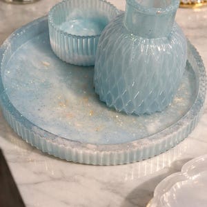 May include: A decorative set comprising a round tray, a small bowl, and a vase, all crafted from light blue resin with a glittery finish. The tray and bowl feature a ribbed design, and the vase has a textured, diamond-like pattern.