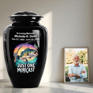 May include: A black cremation urn with a fishing theme. The urn features a colorful graphic of a fish jumping out of the water with a fishing rod and the text "JUST ONE MORE CAST". The urn also includes the text "In Loving Memory Michelle R. Dean Dec 07, 1965 - June 24, 2025".