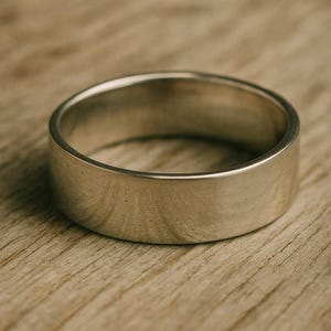 May include: A silver ring with a flat, wide band. The ring is set against a light brown wooden surface. The ring's design is simple and modern, suitable for everyday wear.