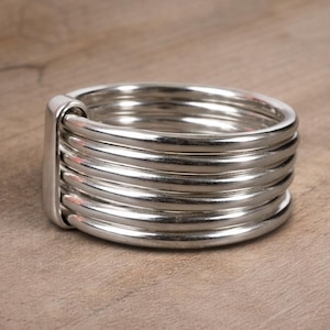 May include: A silver ring featuring multiple stacked bands, with a smooth, polished finish. The ring has a rectangular connecting element. It is presented on a wooden surface.
