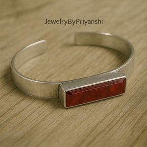 May include: A silver cuff bracelet featuring a rectangular, deep red stone. The bracelet has a simple, modern design. The text "JewelryByPriyanshi" is visible. The bracelet is made of silver.