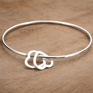 Könnte beinhalten: Ein silbernes Armreif-Armband mit drei herzförmigen Anhängern. Das Armband besteht aus einem dünnen, kreisförmigen Band, wobei die Herzen in der Mitte verbunden sind. Das Armband wird auf einer Holzoberfläche gezeigt.