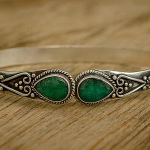 Puede incluir: Brazalete de plata con dos gemas verdes vibrantes en forma de pera. El brazalete presenta un trabajo de plata ornamentado y detallado alrededor de las gemas y a lo largo de la banda. El brazalete se exhibe sobre una superficie de madera.
