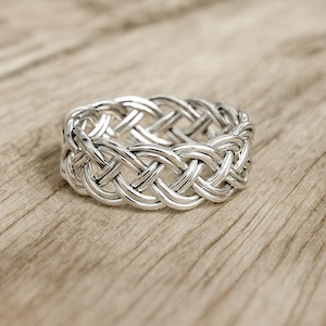 Puede incluir: Un anillo plateado con un diseño trenzado. El anillo tiene una banda gruesa con un intrincado patrón de hebras entrelazadas, creando una apariencia texturizada. El anillo se muestra sobre una superficie de madera de color marrón claro.