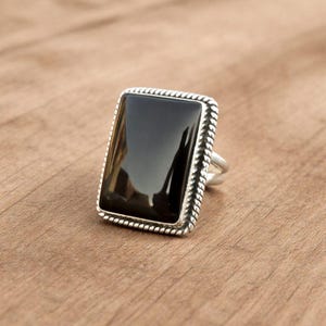 Può includere: Un anello rettangolare in onice nero con una fascia d'argento. La pietra d'onice è incastonata in una cornice d'argento con un bordo a forma di corda. L'anello è esposto su una superficie di legno.