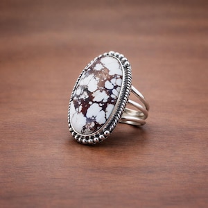 May include: Oval-shaped ring featuring a large, white and brown stone. The stone is set in a silver-colored metal band with a beaded border. The ring has a split shank design, adding a unique touch to this jewelry piece.