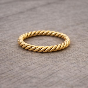 Può includere: Un anello dorato con un design a corda intrecciata. L'anello è circolare e poggia su una superficie grigia. La trama dell'anello è visibile, con ogni torsione che riflette la luce.