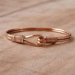 Puede incluir: Una pulsera de color cobre con cierre de gancho y ojo. La pulsera presenta un diseño de doble banda con alambre enrollado a cada lado del cierre. La joya se exhibe sobre una superficie de madera marrón claro.