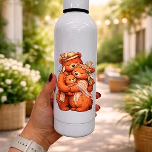 Puede incluir: Botella de agua aislada blanca con un diseño de tres osos abrazándose. Los osos son marrones con detalles florales y de plumas. La botella tiene una tapa blanca y una banda negra. La botella es sostenida por una persona.