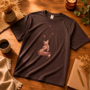May include: A dark brown t-shirt featuring a watercolor illustration of a fox looking up at leaves. The fox is in shades of brown and red. The shirt is short-sleeved and has a crew neck.