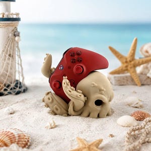 Puede incluir: Un mando de videojuegos rojo descansa sobre una figura de pulpo beige en una playa arenosa. La escena incluye un faro en miniatura, estrellas de mar y conchas, evocando un tema costero. El mando tiene botones negros y un diseño circular.