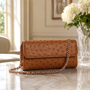 Puede incluir: Un bolso de mano rectangular de color tostado con un patrón texturizado de piel de avestruz, cierre de solapa y correa de hombro de cadena metálica dorada. El bolso tiene una silueta estructurada y un acabado liso que refleja la luz.