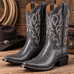 Op de afbeelding: Een paar zwarte cowboylaarzen met een voet van gestructureerd leer met slangenprint en een hoge schacht met gedetailleerd borduurwerk in crème en donkergrijs. De laarzen hebben een spitse neus, een hak en treklussen aan de bovenzijde.