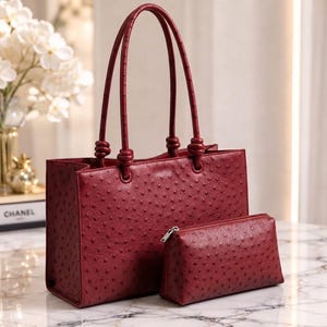 Pode incluir: Uma bolsa tote vermelho-escura e uma bolsa retangular com zíper a combinar, ambas com um acabamento em couro texturizado com relevo de avestruz. A bolsa tote tem duas alças altas e finas com detalhes de nós na base de fixação.