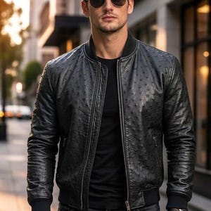 May include: Black leather bomber jacket with a textured pattern, a zip-up front, and ribbed cuffs and hem. The jacket is worn with a black t-shirt and dark wash jeans. The person is wearing sunglasses.