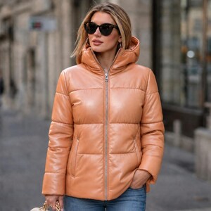 May include: A light orange puffer jacket with a hood and a silver zipper. The jacket has a quilted design and is worn by a person wearing sunglasses.