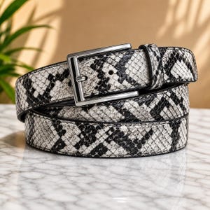 Op de afbeelding: Een riem met een zwart-wit slangenleerpatroon en een gepolijste zilverkleurige metalen gesp. De riem is opgerold en toont de textuur van de reptielenprint over de hele band, inclusief een metalen lus om het uiteinde vast te zetten.