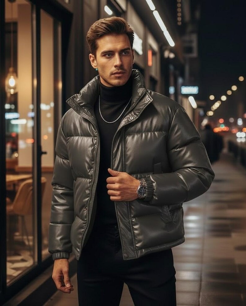 May include: A dark gray puffer jacket with a high collar and a shiny finish. The jacket is worn over a black turtleneck and paired with black pants. A silver chain and watch complete the look. The background shows a city street at night.