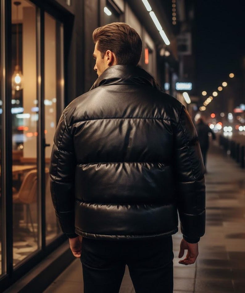 May include: A black puffer jacket with a shiny, leather-like appearance. The jacket has horizontal stitching and a high collar. The person is walking on a sidewalk at night, with storefronts and streetlights in the background.