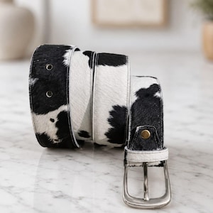 May include: A belt featuring a black and white cow print pattern with a textured hair-on-hide finish. The belt includes a silver-toned metal buckle, a single metal rivet near the buckle, and multiple circular holes along the strap for adjustment.