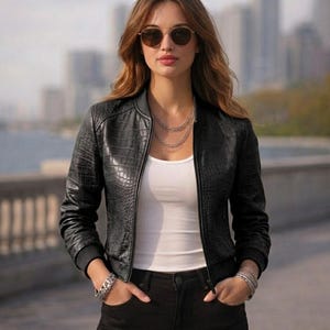 May include: A black faux leather bomber jacket with a crocodile texture. The jacket has a ribbed collar and cuffs. The person is wearing a white tank top, black jeans, and sunglasses. Multiple silver bracelets and necklaces complete the look.