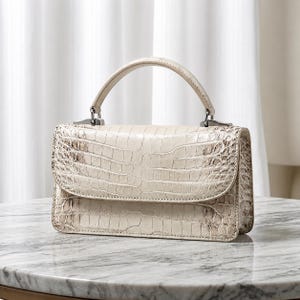 Peut inclure: Un sac à main structuré de couleur crème blanc cassé avec un motif texturé en peau de crocodile. Le sac présente une poignée supérieure unique, une silhouette rectangulaire et une fermeture à rabat avec des attaches en métal argenté.