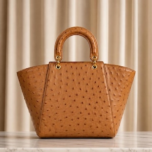 May include: A tan handbag with a distinct textured surface featuring small, evenly spaced indentations. The bag has a structured, trapezoidal shape, a single rounded top handle, and gold-toned metal hardware connecting the handle to the body.