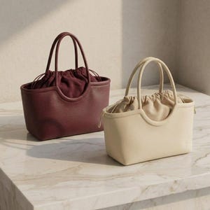Pode incluir: Duas bolsas estruturadas com alças duplas superiores e fecho de tecido com cordão. Uma bolsa é bordô escuro e a outra é creme, ambas com acabamento exterior texturizado e um design de abertura superior curva.