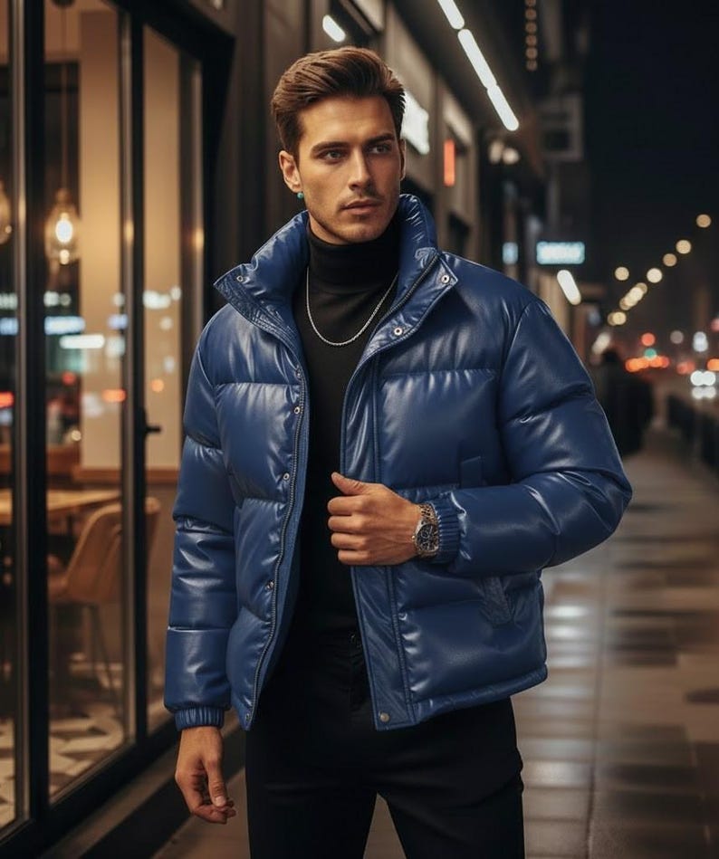 May include: A blue puffer jacket with a high collar. The jacket has a shiny, leather-like appearance and a zipper closure. The person is wearing a black turtleneck and a silver chain.