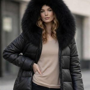 Può includere: Giacca trapuntata nera con un grande cappuccio bordato di pelliccia nera e soffice. La giacca è indossata sopra un top color beige chiaro. La modella indossa jeans blu scuro. La giacca ha un design trapuntato e una chiusura con cerniera.