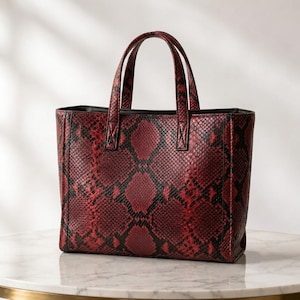 May include: A structured tote bag featuring a bold red and black snakeskin pattern. The handbag has two top handles, a rectangular silhouette, and a black interior lining visible at the top edge. The exterior surface displays a textured scale design.