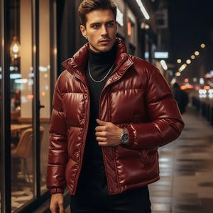 May include: A burgundy puffer jacket with a high collar. The jacket has a shiny, leather-like appearance and is buttoned up. The person is wearing a black turtleneck and a silver chain. A watch is visible on the wrist.