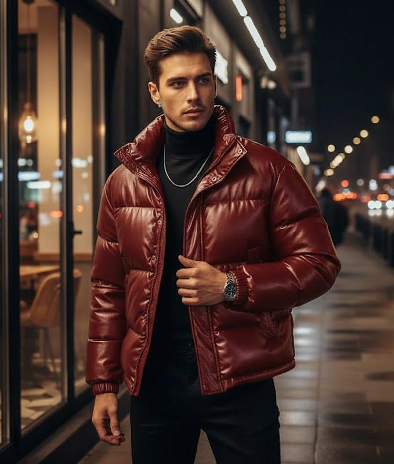 May include: A burgundy puffer jacket with a high collar. The jacket has a shiny, leather-like appearance and is buttoned up. The person is wearing a black turtleneck and a silver chain. A watch is visible on the wrist.