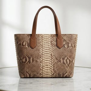 May include: A structured tote bag featuring a textured snakeskin pattern in shades of beige, tan, and brown. The bag has two solid brown leather handles attached with stitched tabs. The design includes a vertical panel down the center.