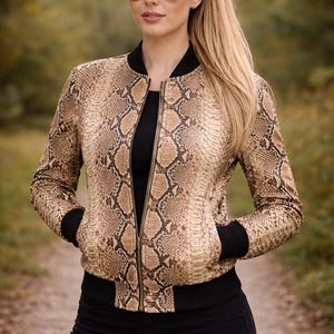 May include: A brown and tan snakeskin-patterned bomber jacket with black trim and a zipper closure. The jacket is worn over a black top and paired with black pants. The model is wearing sunglasses and has blonde hair pulled back in a ponytail.