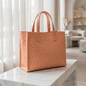Puede incluir: Un bolso tote de color melocotón con un patrón texturizado de piel de avestruz. El bolso presenta una silueta rectangular, dos asas verticales superiores y una base estructurada. La superficie tiene un aspecto punteado uniforme.