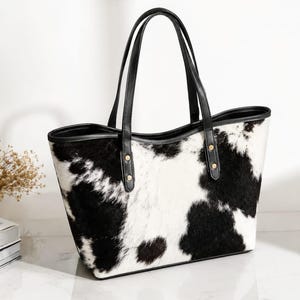 Puede incluir: Un bolso tote con un patrón de manchas de vaca en blanco y negro y una textura similar al pelaje. El bolso incluye dos correas negras para el hombro sujetas con remaches de metal dorado y un borde negro a lo largo de la parte superior.