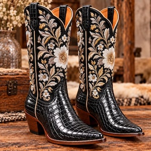 Könnte beinhalten: Ein Paar schwarze Stiefel mit einem glänzenden, strukturierten Krokodilledermuster am Fuß und an der Ferse. Die hohen Schäfte bestehen aus schwarzem Material, das mit aufwendigen, mehrfarbigen Blumenstickereien in Weiß, Gold und Beige verziert ist.