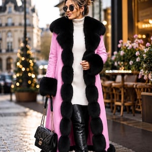 Könnte beinhalten: Ein langer rosa Mantel mit auffälligem schwarzem Pelzkragen, Ärmelbündchen und Frontbesatz. Der Mantel wird über einem weißen gerippten Rollkragenpullover und schwarzen Overknee-Stiefeln aus Leder getragen. Eine schwarze gesteppte Handtasche mit Goldkette.