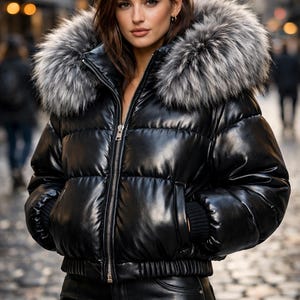 May include: A black puffer jacket with a faux fur hood. The jacket has a shiny, leather-like finish and a silver zip. The model is wearing black leather trousers. The background is a blurred city street.