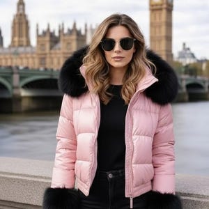 Puede incluir: Chaqueta acolchada rosa claro con cuello y puños de piel negra. Se lleva con vaqueros negros y una camiseta negra. El fondo muestra un río y edificios.