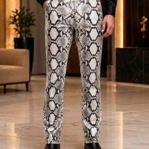 May include: Full-length trousers featuring a white and black snakeskin pattern. The pants have a straight-leg cut, a standard waistband with belt loops, and a black belt. The design includes a front button closure and side pockets.