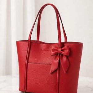 Puede incluir: Un bolso tote de color rojo brillante con una superficie texturizada y granulada. El bolso cuenta con dos correas verticales, dos asas superiores y un lazo decorativo fijado en la parte superior derecha. El diseño es estructurado con base plana.