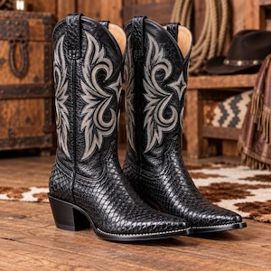 Puede incluir: Un par de botas vaqueras negras con la parte inferior texturizada en patrón de piel de serpiente y una caña de cuero liso. La caña está decorada con intrincados bordados de color gris plateado en forma de espiral y tiene tiradores.