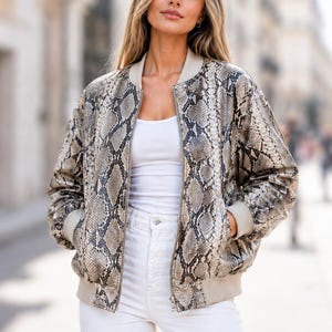 Puede incluir: Chaqueta bomber con estampado de piel de serpiente en tonos grises y beige. La chaqueta tiene cuello, puños y dobladillo de canalé, y cierre de cremallera frontal. La modelo lleva una camiseta sin mangas blanca y vaqueros blancos.