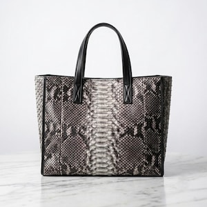 May include: A structured tote bag featuring a grey and black snakeskin pattern with a textured scale finish. The bag includes two solid black leather top handles and black leather trim along the vertical side panels and the top opening edge.