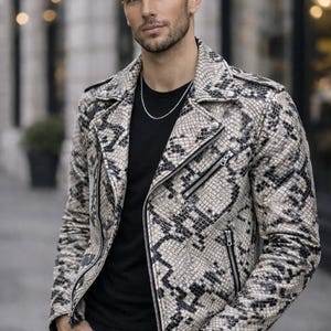 Może przedstawiać: Skórzana kurtka ze wzorem skóry węża w odcieniach beżu, czerni i szarości. Kurtka w stylu motocyklowym ma kołnierz z klapami, asymetryczne zapięcie na zamek i wiele kieszeni zapinanych na zamek. Model ma na sobie czarny t-shirt i czarne dżinsy.