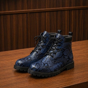 May include: A pair of blue and black snake-skin patterned boots with black laces and a black sole. The boots are made of a shiny material and are sitting on a wooden surface.