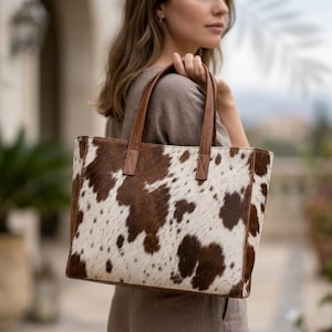May include: A rectangular tote bag featuring a brown and white cowhide pattern with a textured fur-like appearance. The bag includes two brown leather shoulder straps attached to the top edge, providing a structured shape for carrying items.
