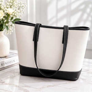 May include: A structured tote bag featuring a white pebbled body with a solid black base and black trim. The bag includes two long black shoulder straps with visible white stitching, a wide top opening, and a clean, minimalist rectangular silhouette.